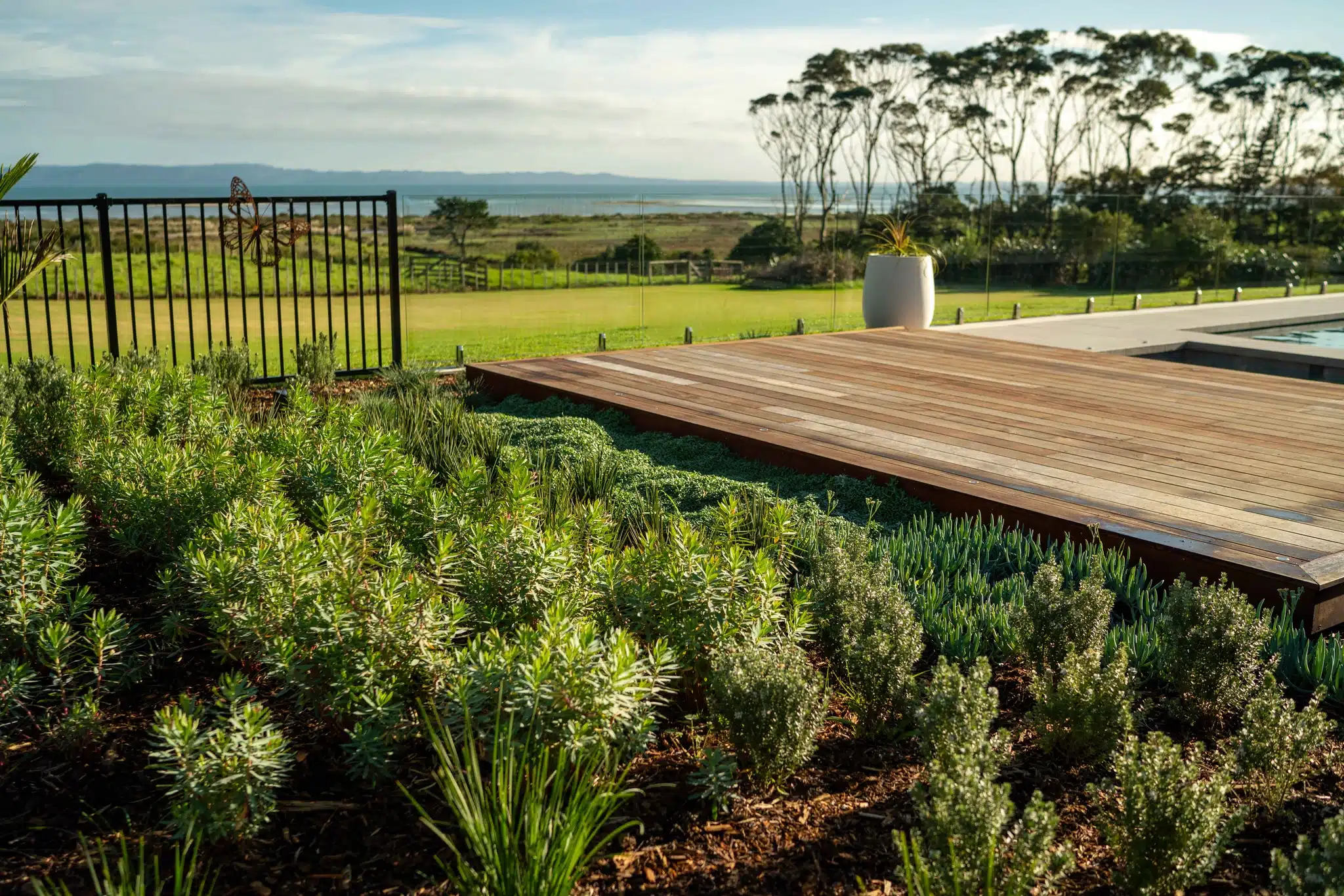 Scenic garden landscaping with a rock wall, diverse plants, wooden deck, and a fence overlooking rural farmland and greenhouses.
