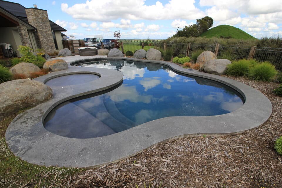 Modern outdoor swimming pool with an integrated, bubbling spa. Features include sleek patio pavers and elegant backyard landscaping.