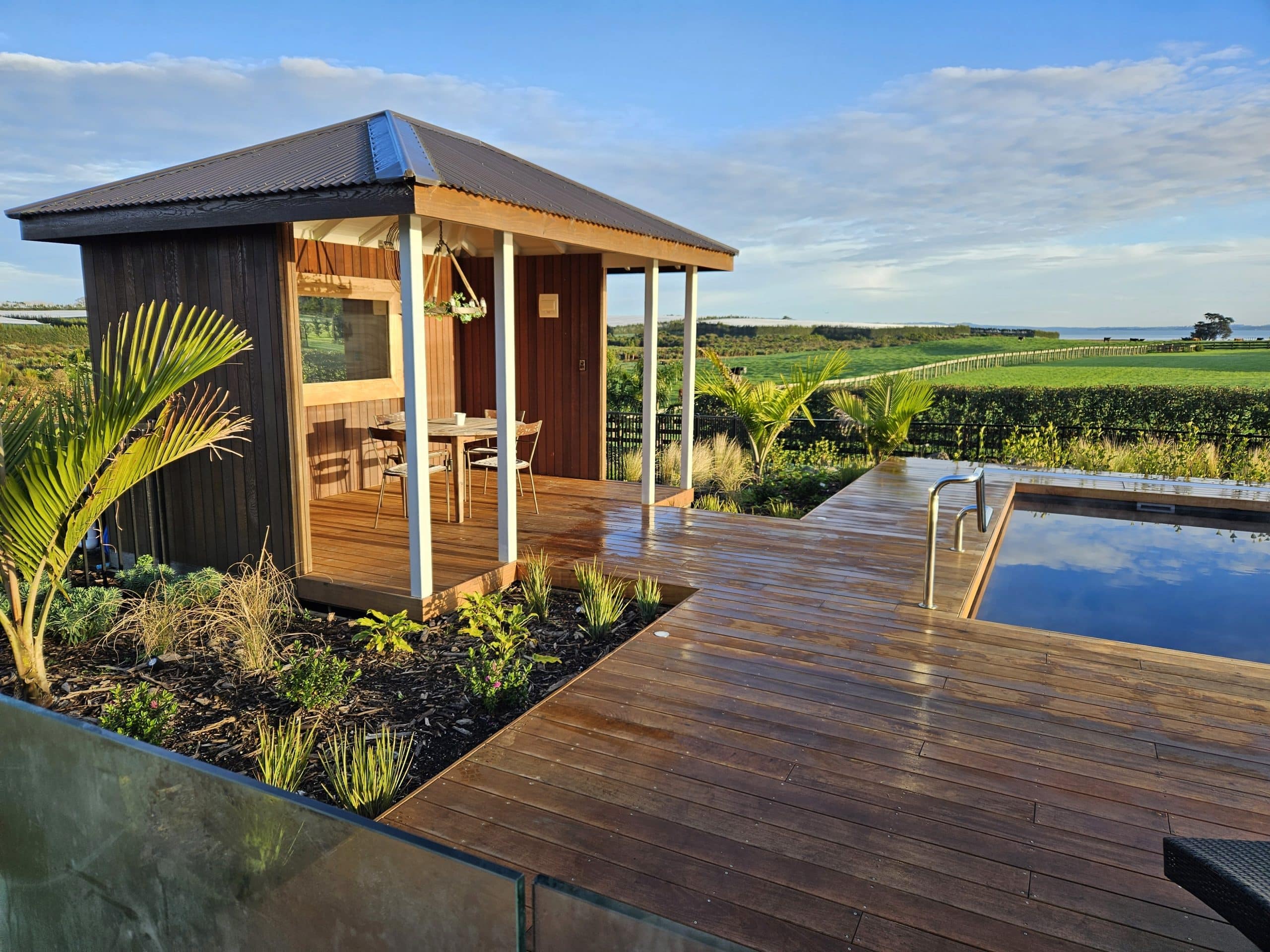 Scenic garden landscaping with a rock wall, diverse plants, wooden deck, and a fence overlooking rural farmland and greenhouses.